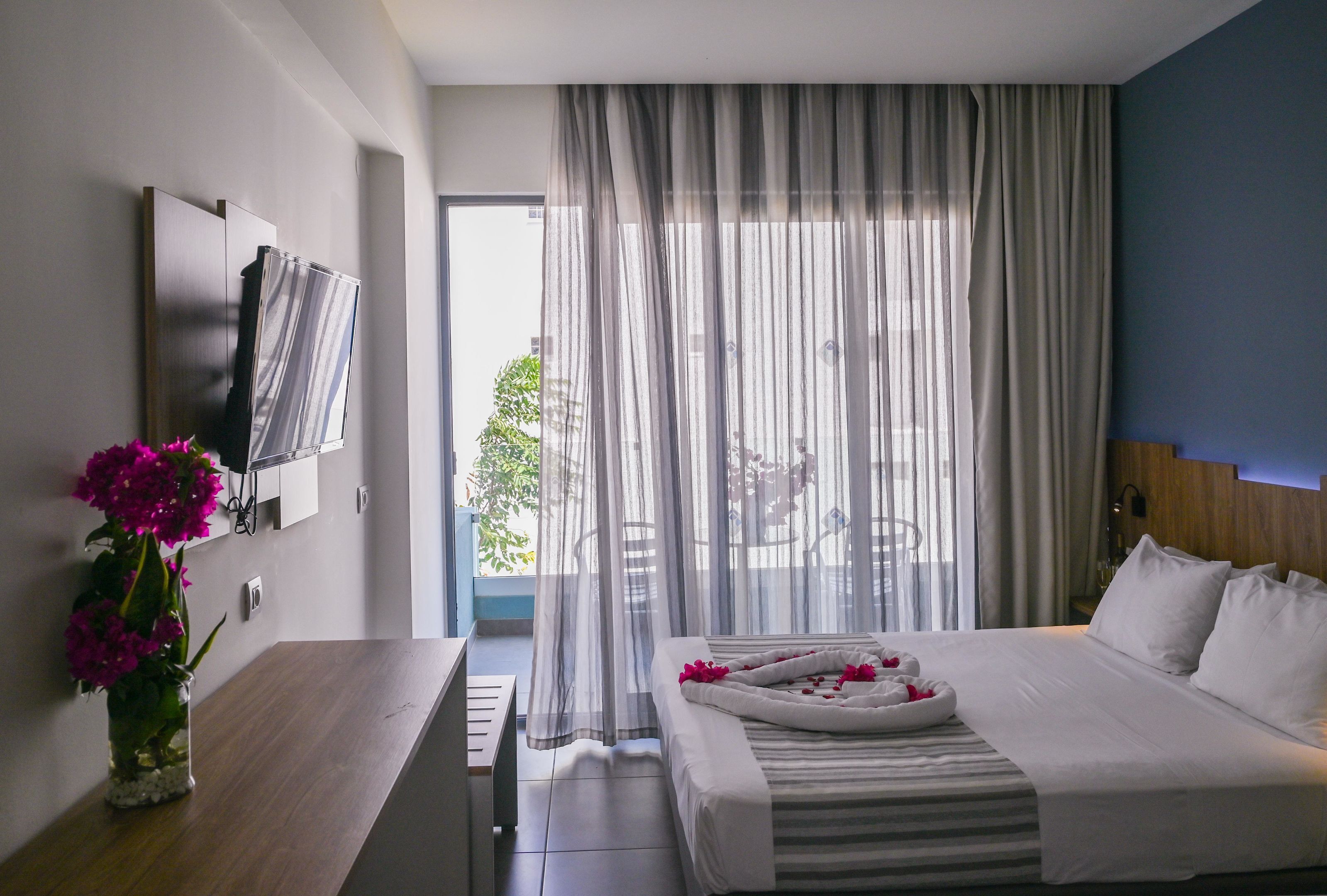 A hotel room with a bed, a TV mounted on the wall, a small table with a vase of flowers, and a view of the outdoors through sheer curtains.