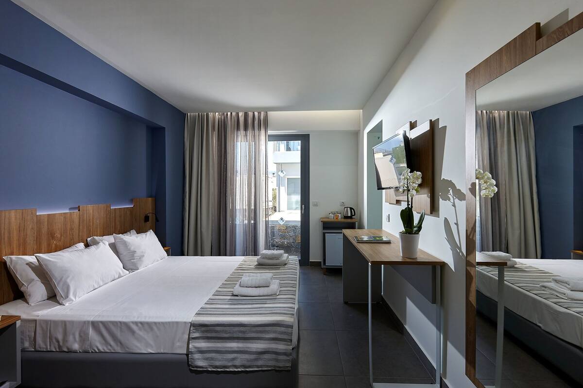 A hotel room with a large bed, a desk, a mirror, and a view of the outside.