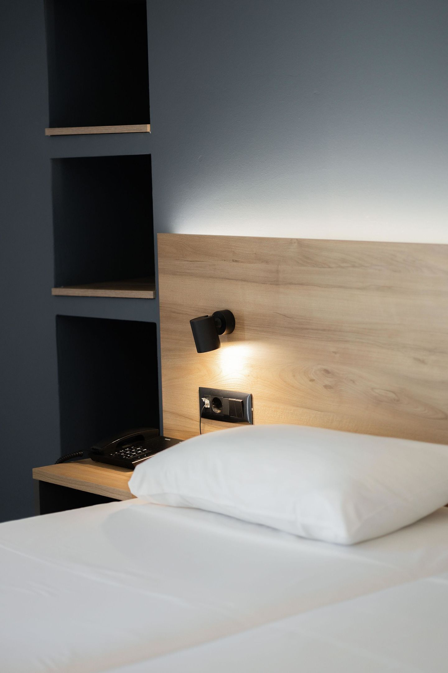 Habitación de hotel con cabecera de madera, mesita de noche con teléfono negro, almohada blanca y luz fijada en la pared.