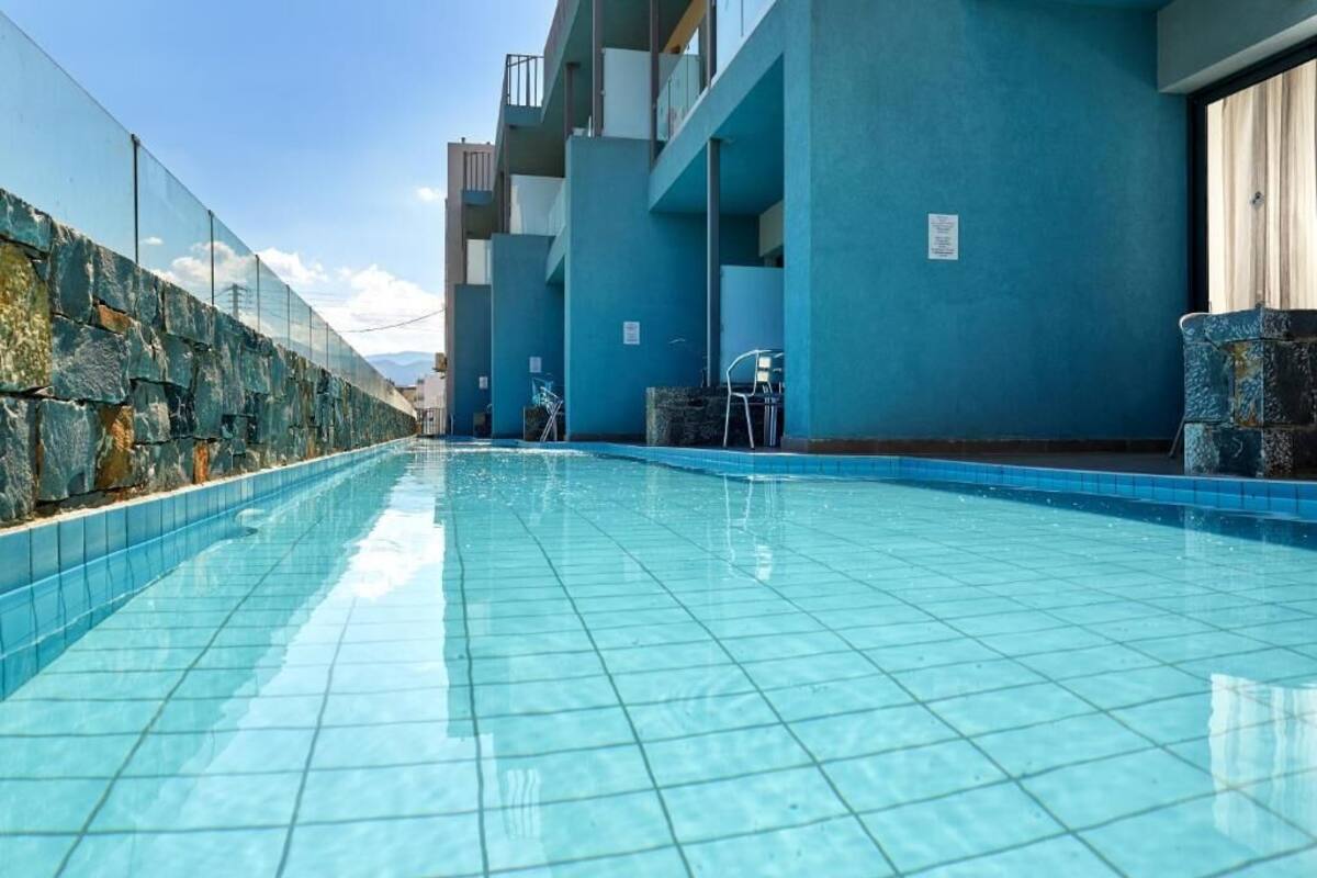 A swimming pool with blue tiled flooring, surrounded by a stone wall and a blue building with balconies.