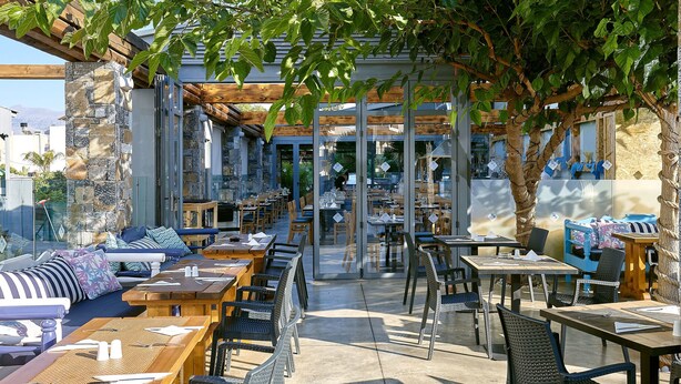 Un café al aire libre con mesas y sillas de madera, pilares de piedra y una pérgola cubierta de plantas trepadoras.