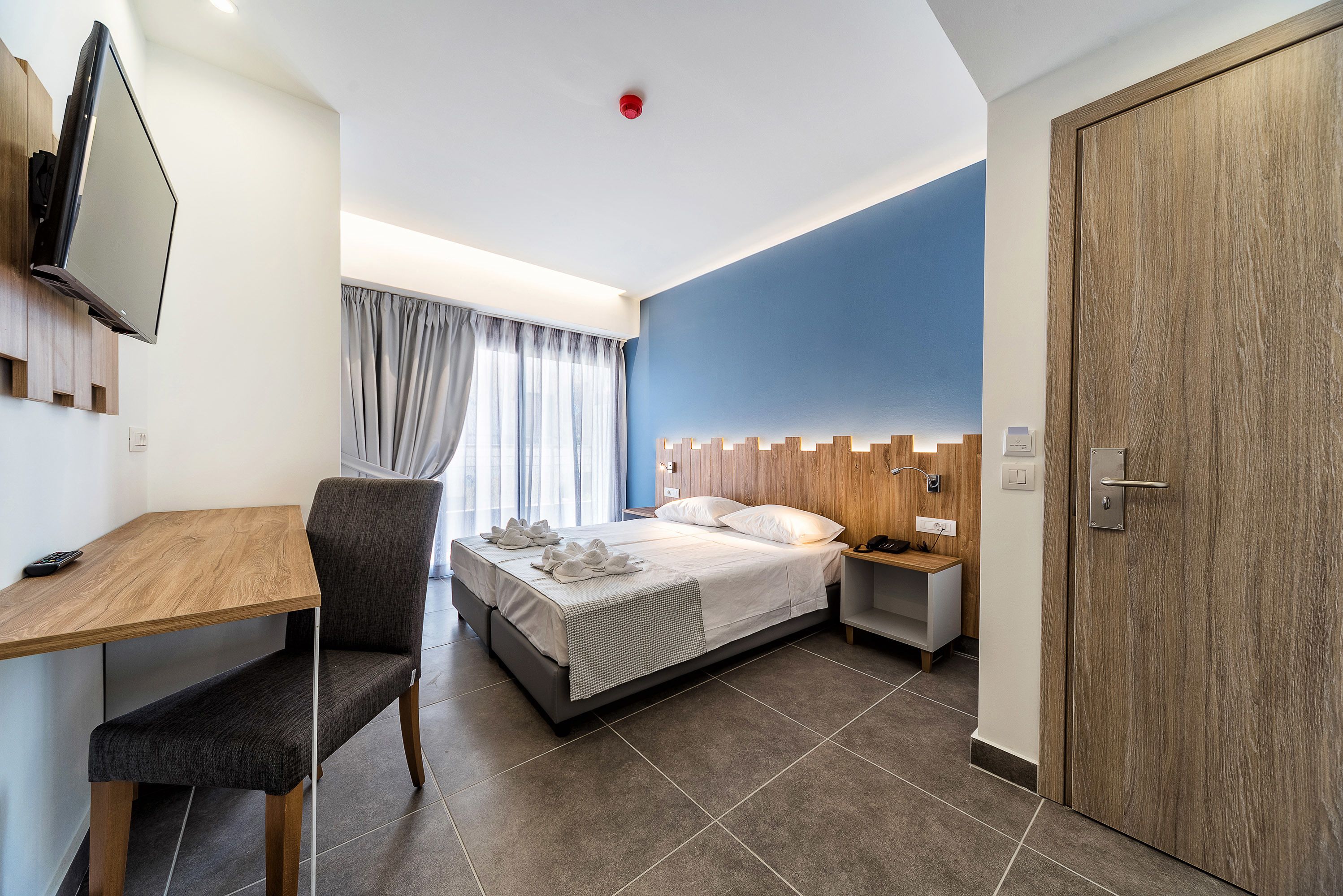A modern hotel room with a bed, a desk, a chair, a TV, and a wardrobe.