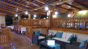 Terraza interior de madera con asientos y mesas, un balcón con barandilla y vistas al exterior.