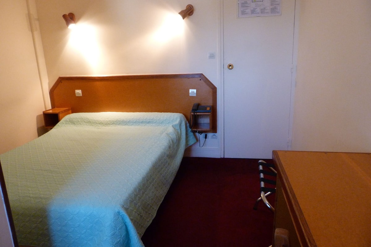 Une petite chambre avec un lit simple, une tête de lit en bois, un téléphone sur la table de chevet et une moquette rouge.
