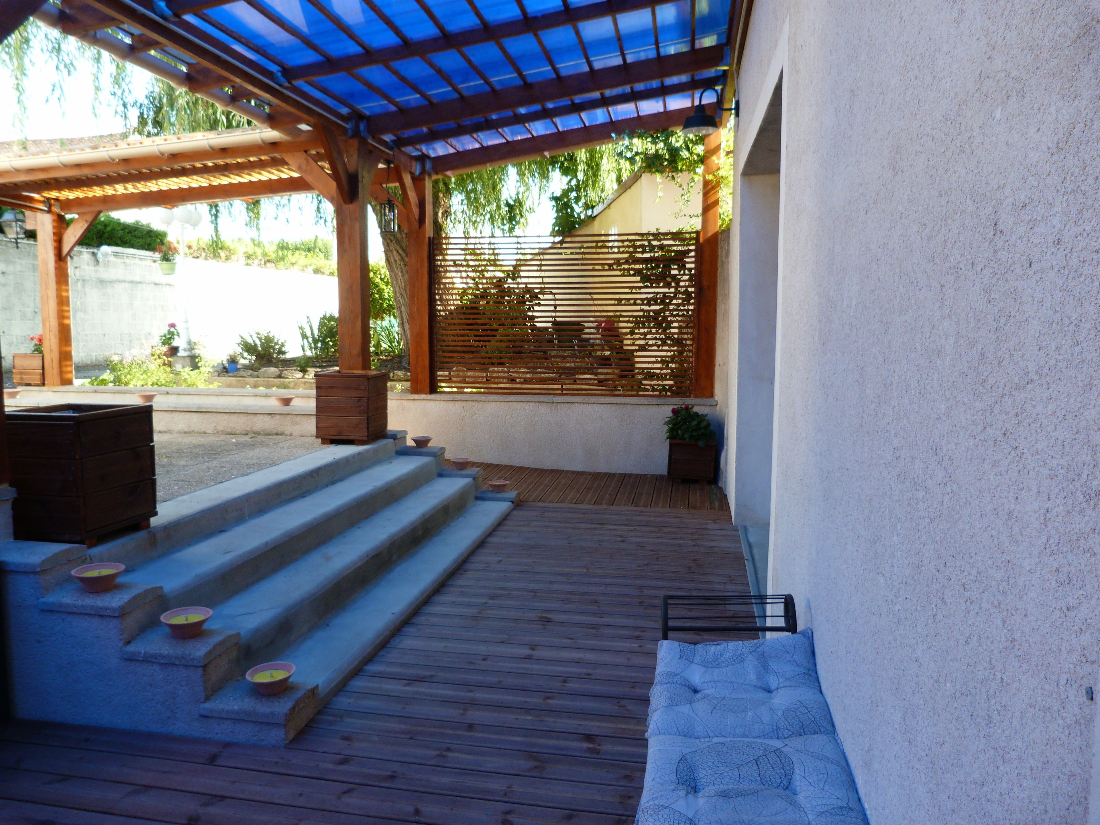 Un espacio al aire libre cubierto con escalones de madera que conducen a una zona de estar, una pérgola con techo azul y una valla de madera con plantas trepadoras.