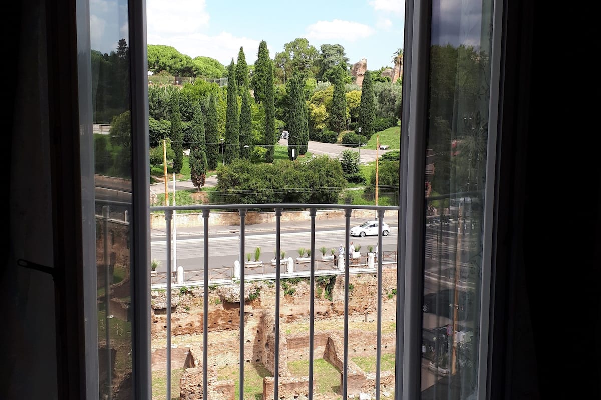 A room with a large sliding glass door leading to an outdoor area with a view of a park, a road, and some ruins.