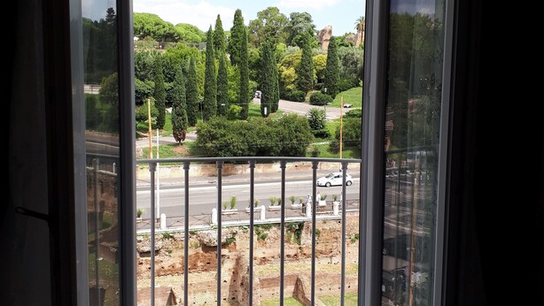 A room with a large sliding glass door leading to an outdoor area with a view of a park, a road, and some ruins. - Colosseo Rooms Imperial Rome (Rome)