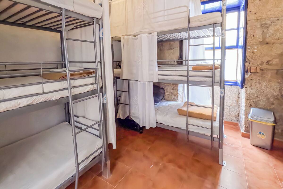 A dormitory room with bunk beds, a window with blue trim, and a trash bin.