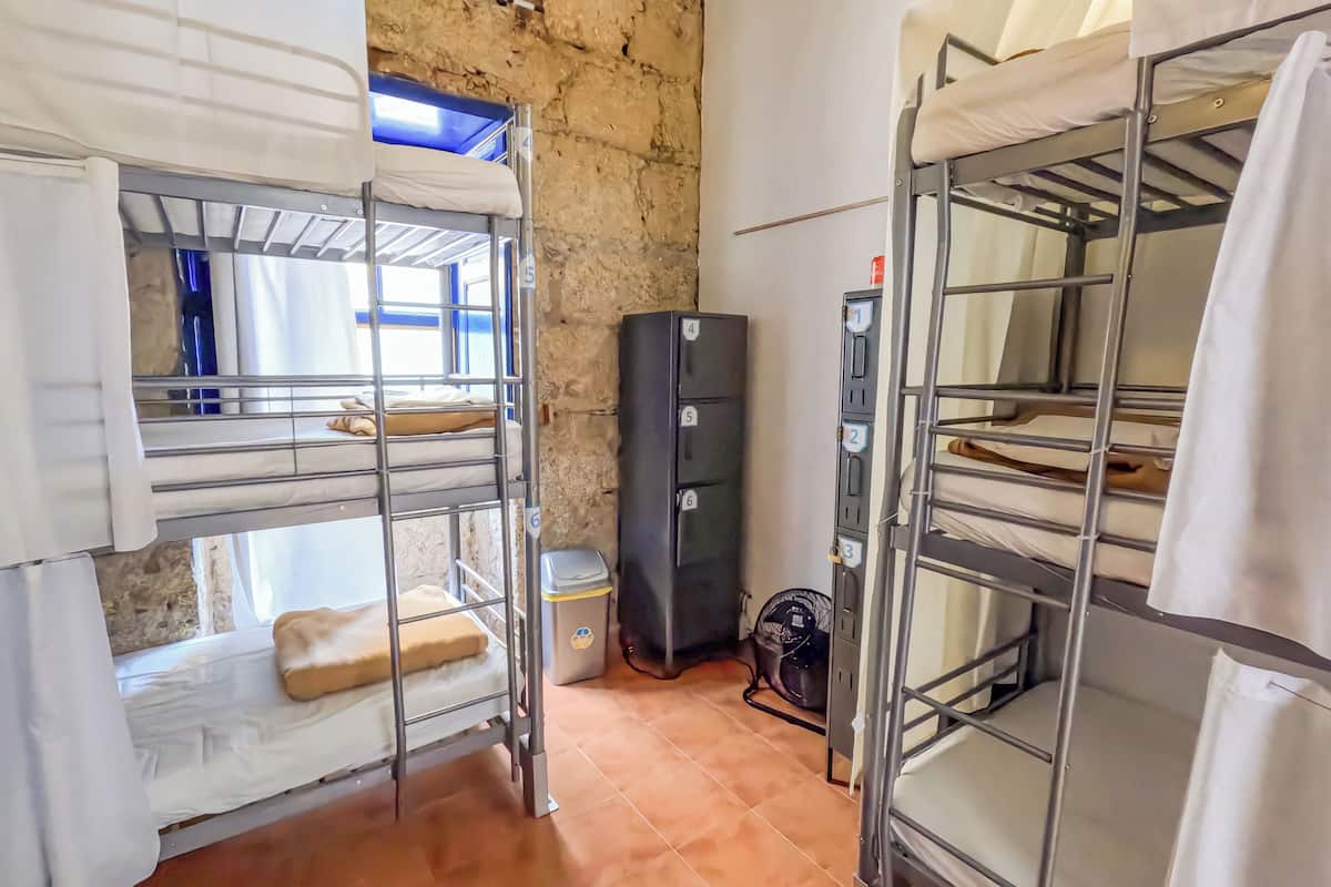 A dormitory room with bunk beds, a black refrigerator, a trash bin, and a heater.