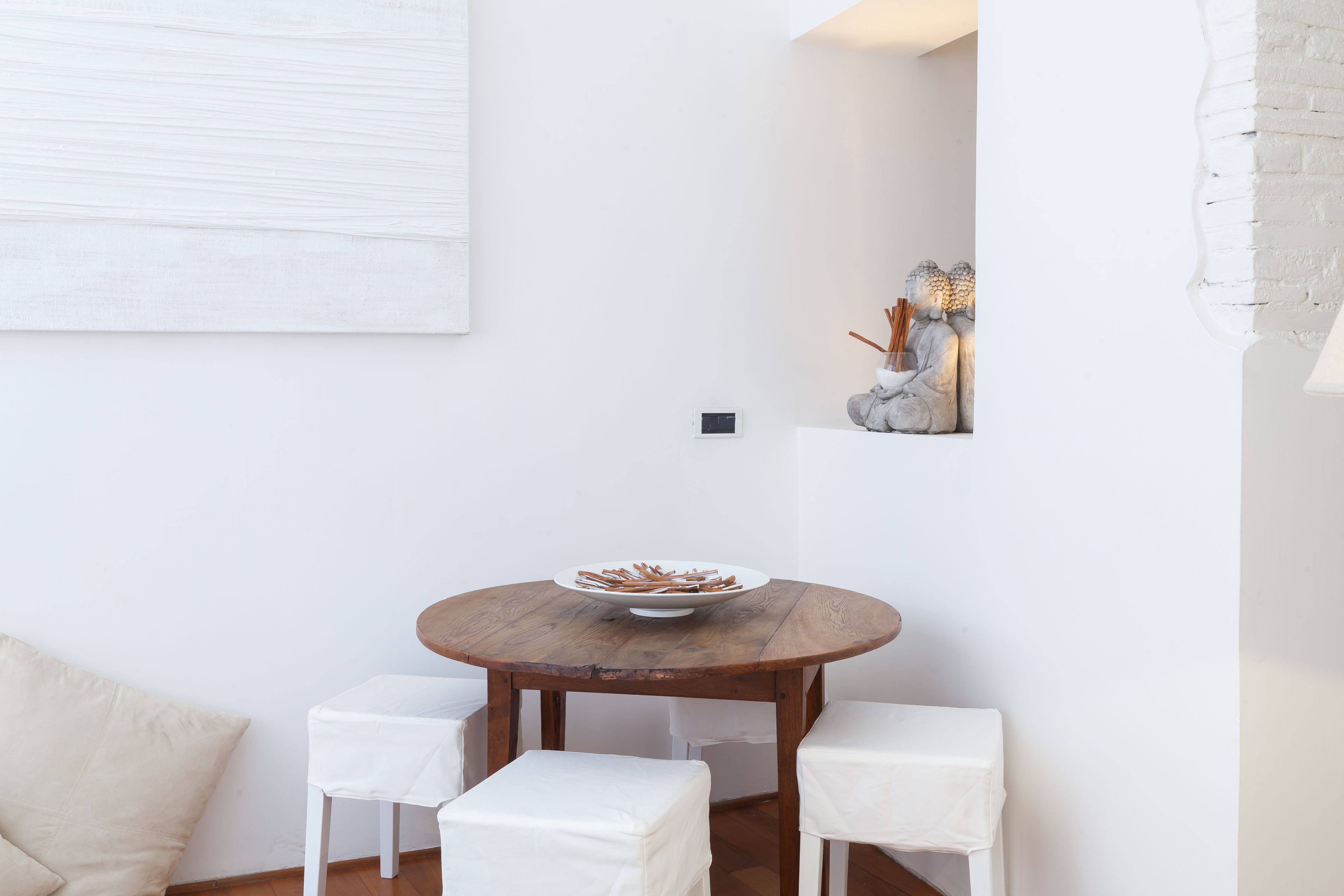 Una mesa redonda de madera con un bol de aperitivos, dos sillas blancas y un banco blanco acolchado.