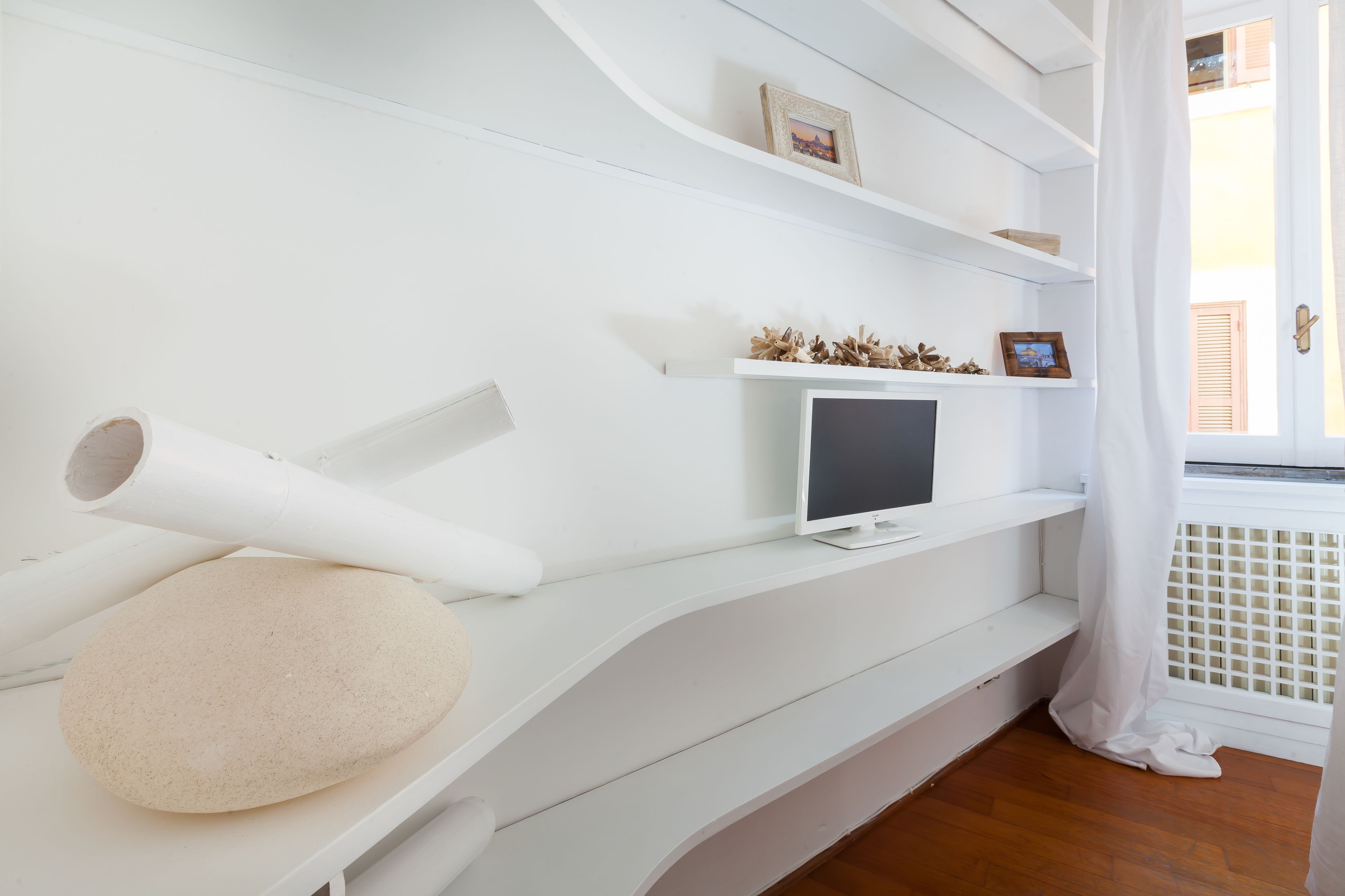 A modern room with white walls, a flat-screen TV on a shelf, and a window with a white curtain.