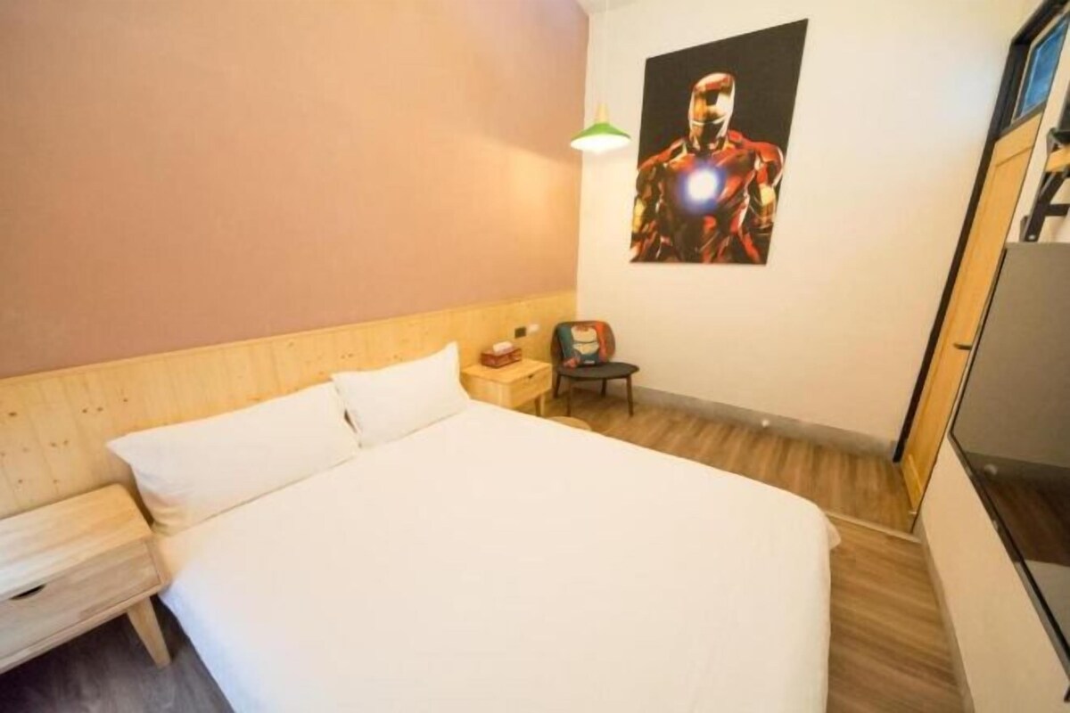 A hotel room with a wooden headboard, a bed with white linens, a small bedside table, a nightstand, a green wall lamp, and a poster of a superhero on the wall.