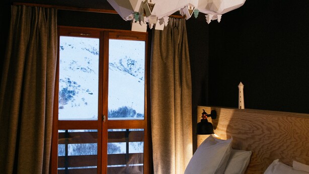 A hotel room with a large bed, a wooden headboard, a window with a view of snow-covered mountains, and a ceiling with a geometric light fixture. - Ho36 Les Menuires (Les Belleville)