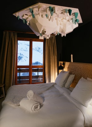 A hotel room with a large bed, a wooden headboard, a window with a view of snow-covered mountains, and a ceiling with a geometric light fixture. - Ho36 Les Menuires (Les Belleville)