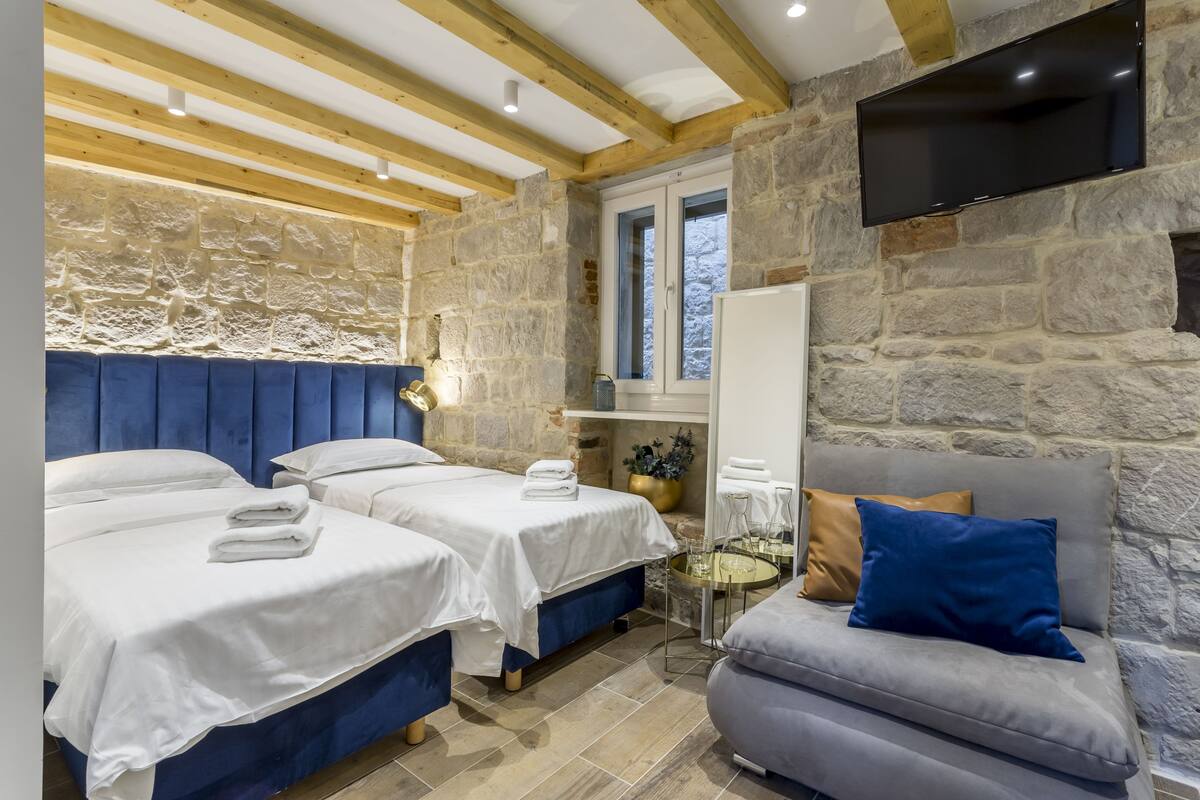 A bedroom with two beds, a sofa, stone walls, wooden beams, a TV, and a window.