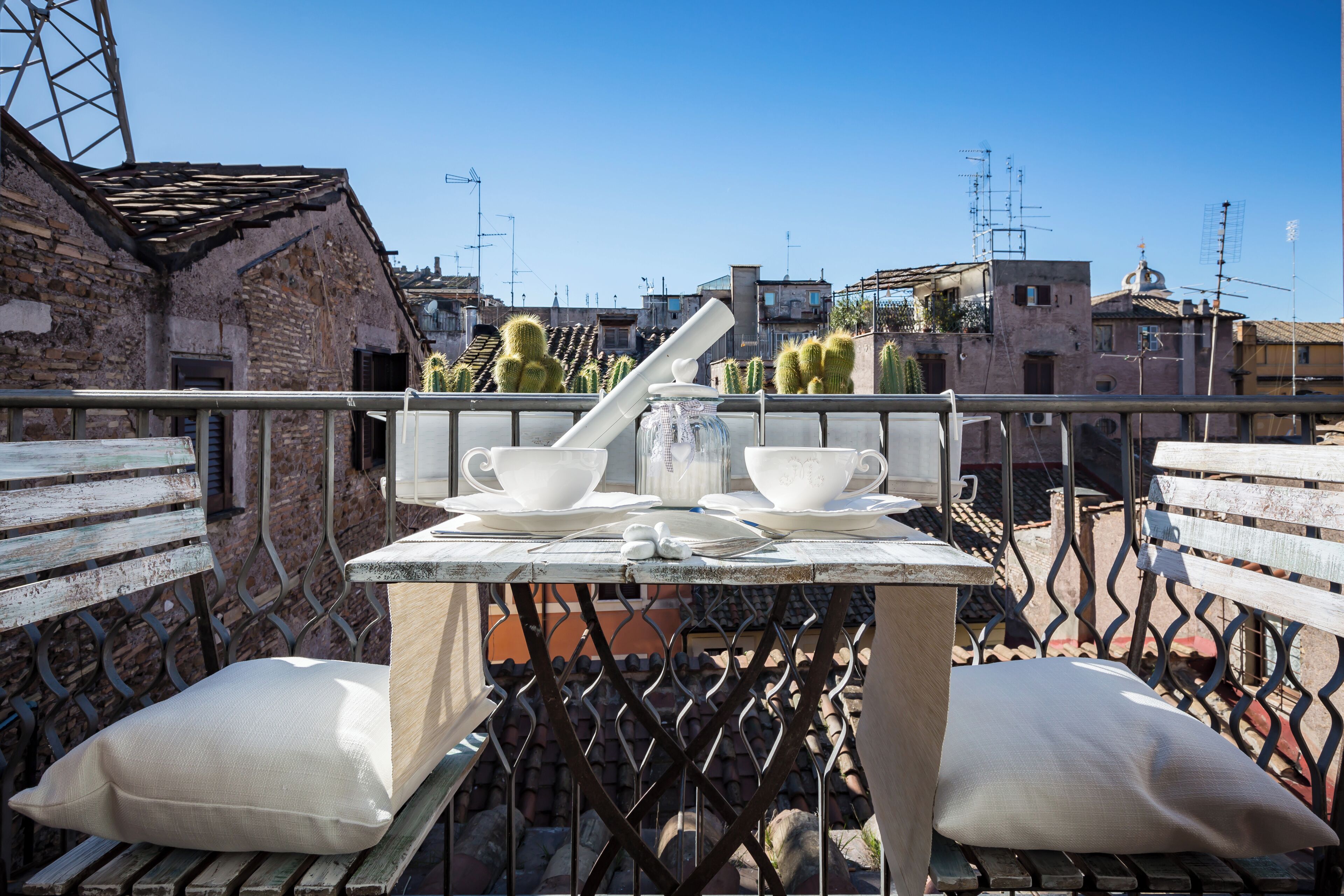 A rooftop terrace with a table set for two, featuring white cups and saucers, a glass jar, and a metal spoon. The terrace has a metal railing and is surrounded by buildings with visible antennas.