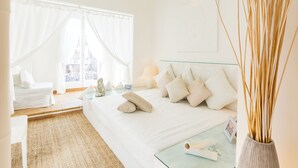 A modern bedroom with a large bed, a glass bedside table, and a view of a cityscape through sheer curtains.