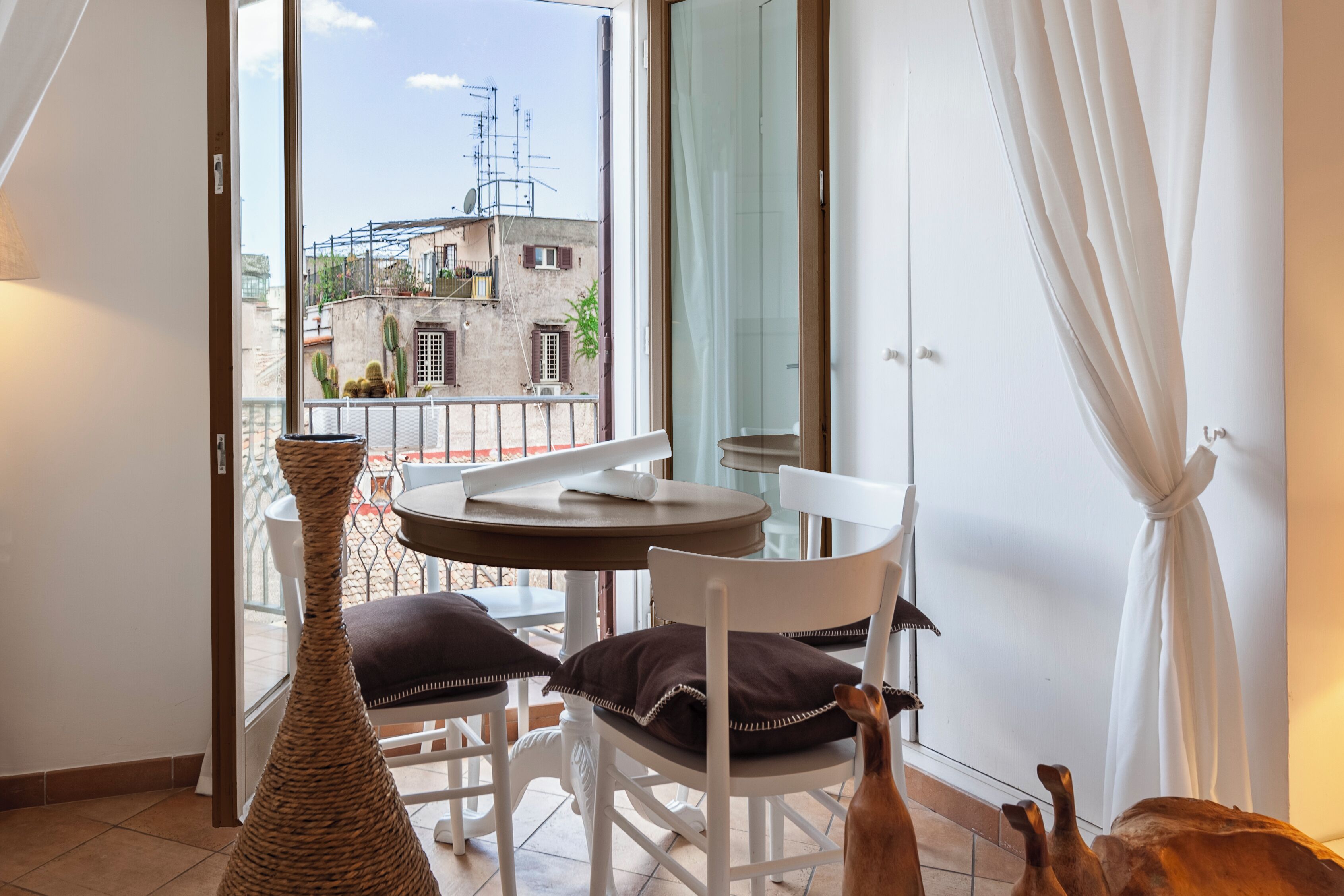 A balcony with a table and chairs, a wicker vase, and a view of buildings and a satellite dish.