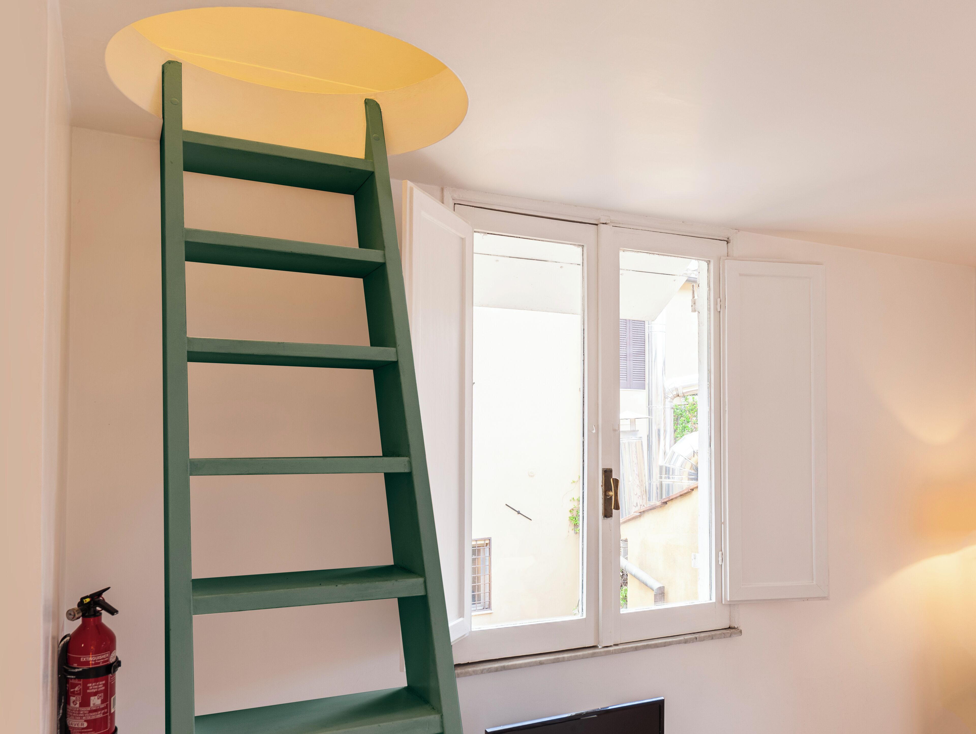 A room with a green ladder leaning against a wall, a white door, and a window with shutters.
