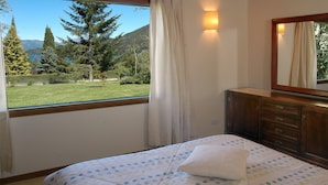 Een slaapkamer met een bed, een dressoir, een spiegel en een raam met uitzicht op een bos en bergen.