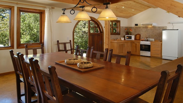 Een eetgedeelte met een houten tafel en stoelen, een keuken op de achtergrond en grote ramen met gordijnen.