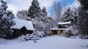Ein mit Schnee bedecktes Chalet mit Holzfassade und gelbem Anstrich, umgeben von schneebedeckten Bäumen und blauem Himmel.