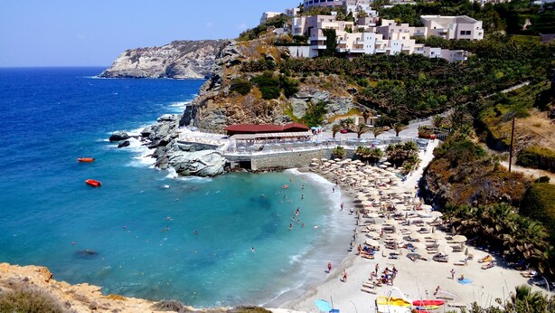 A coastal beach with a rocky shoreline, a sandy beach area with umbrellas and lounge chairs, and buildings on a hillside. - Experience & Enjoy Sea View Studios (Malevizi)