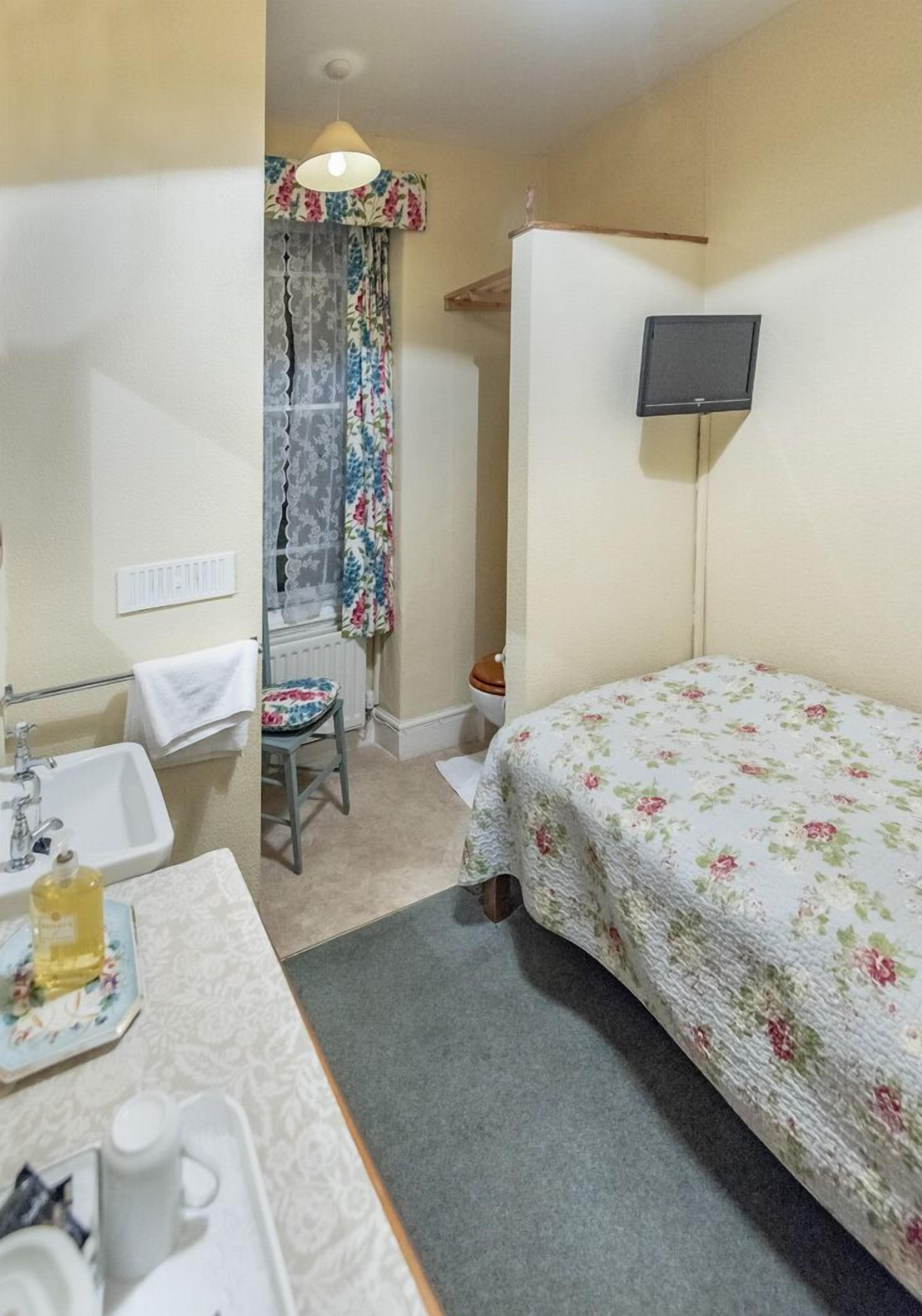 Ein kleines, gemütliches Zimmer mit einem Einzelbett, einem Nachttisch, einem Sessel, einem Waschbecken und einem an der Wand befestigten Flachbildfernseher.