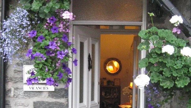 Une entrée en pierre, avec une porte, un panneau indiquant les places disponibles et divers pots de fleurs.