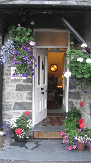 Entrada de un edificio de piedra con una puerta, un cartel que indica habitaciones disponibles y varias macetas con flores.