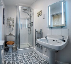 Un baño con cabina de ducha de vidrio, inodoro con asiento de madera, lavamanos con espejo superior y baldosas de suelo estampadas.