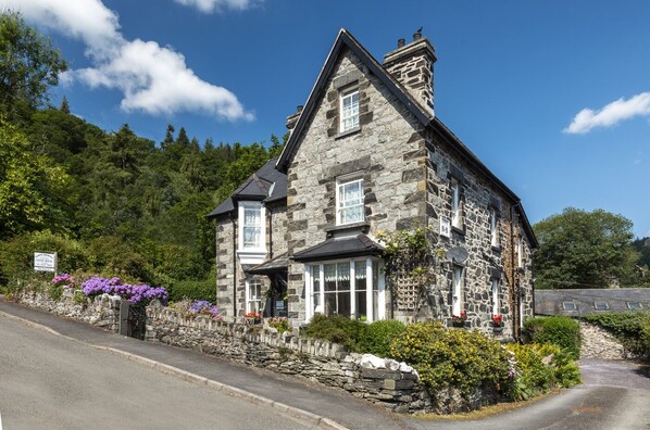 A stone building with a pitched roof, a sign indicating it's a guest house, and a stone wall with flowers.