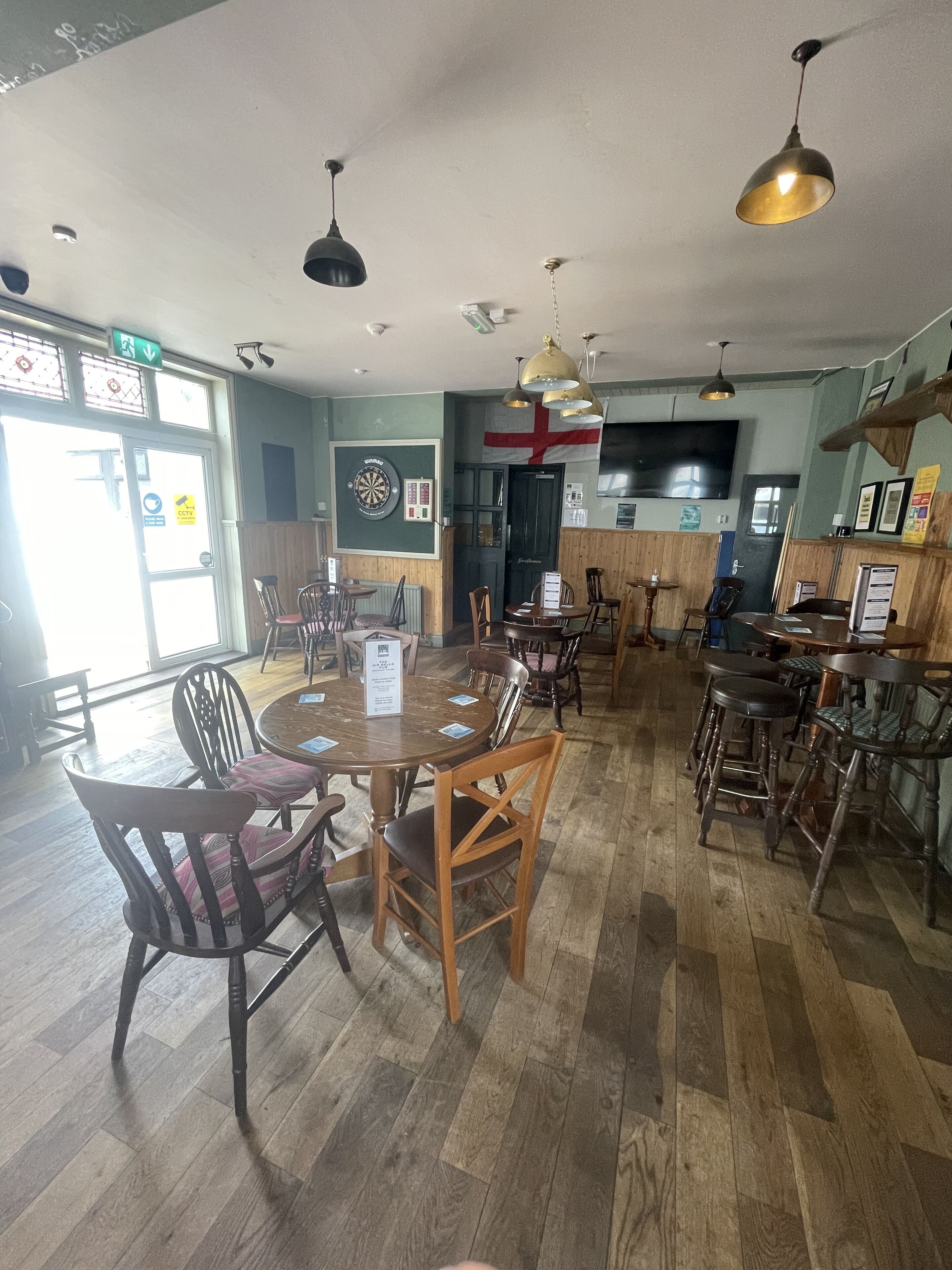 Interno di un ristorante con pavimento in legno, tavoli rotondi e sedie. Sono presenti luci a sospensione e un grande televisore appeso al muro.