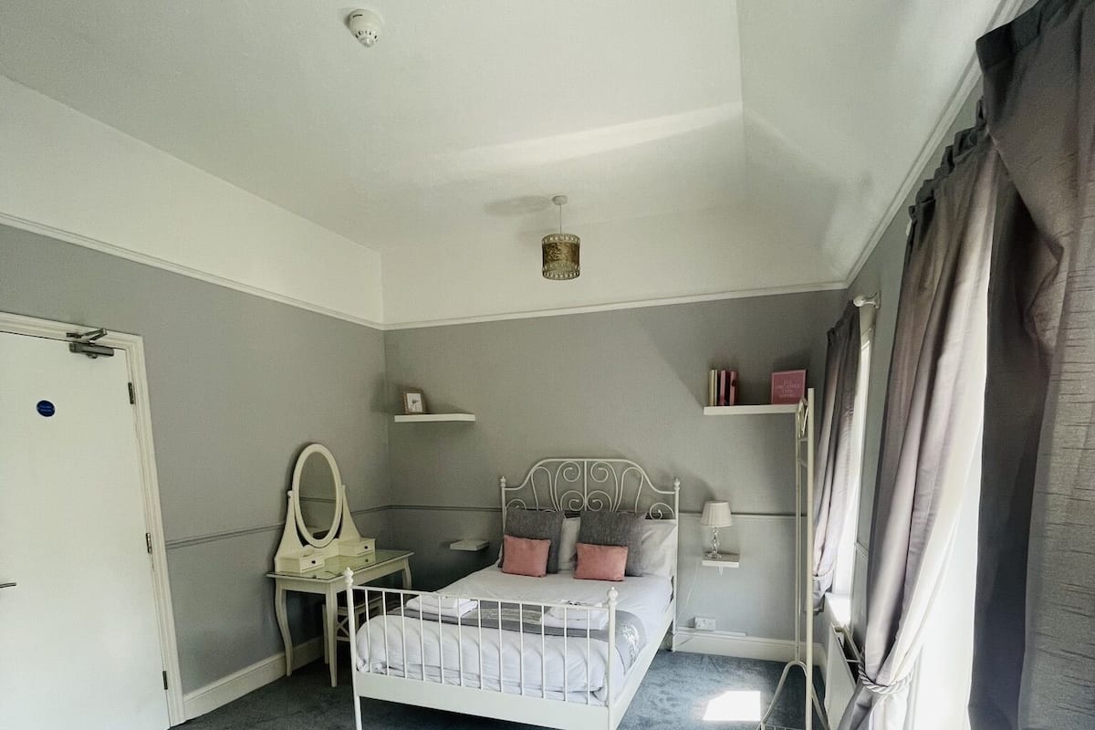 A bedroom with a single bed, a bedside table, a mirror, a lamp, and a window with curtains.