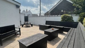Un'area patio esterna con panche e tavoli in legno nero, un muro di mattoni bianchi e vista su un edificio di fronte.