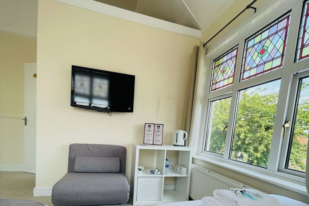 A hotel room with a bed, a gray armchair, a television, a white cabinet, and a window with stained glass.