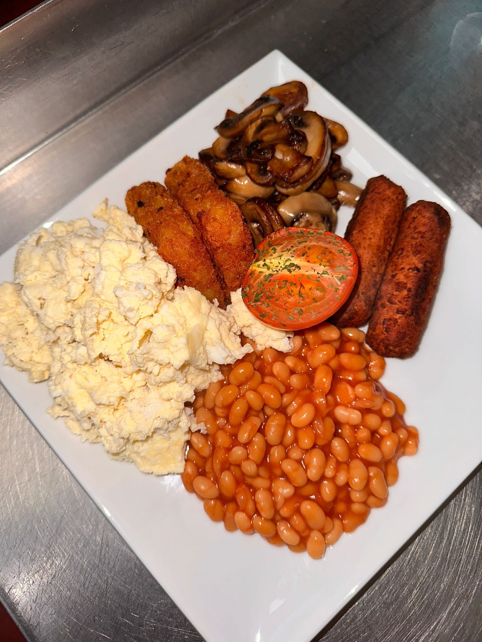 A plate of food including scrambled eggs, baked beans, mushrooms, sausages, and a slice of tomato.