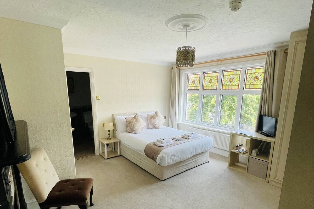 A bedroom with a bed, a chair, a nightstand, a TV, and a window with stained glass.