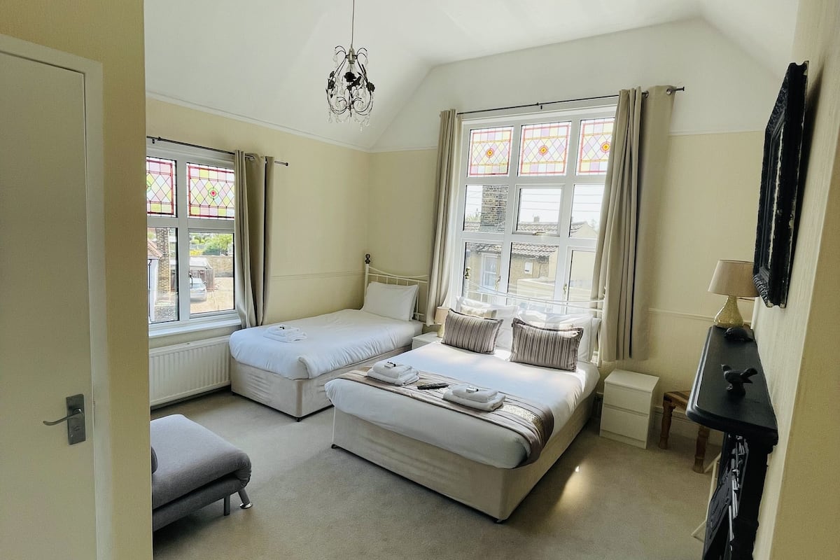 A bedroom with two beds, a window with curtains, a radiator, a hanging light fixture, and a small table.