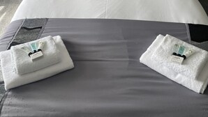 Un letto rifatto con cura, lenzuola grigie e diversi cuscini, un comodino bianco con una lampada e una presa elettrica sulla parete.
