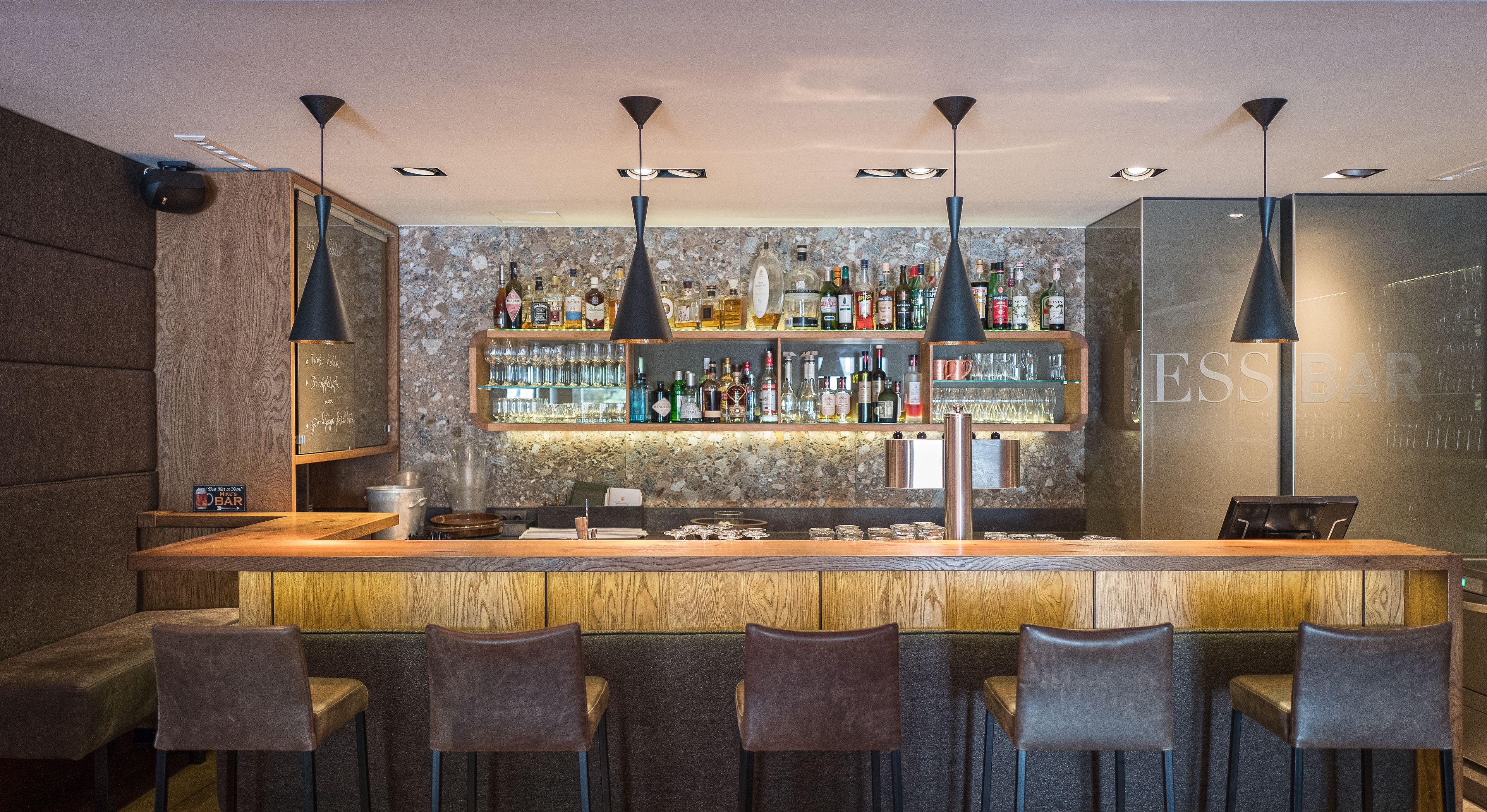 A bar area with a wooden counter, multiple bar stools, and a variety of liquor bottles and glasses on shelves.