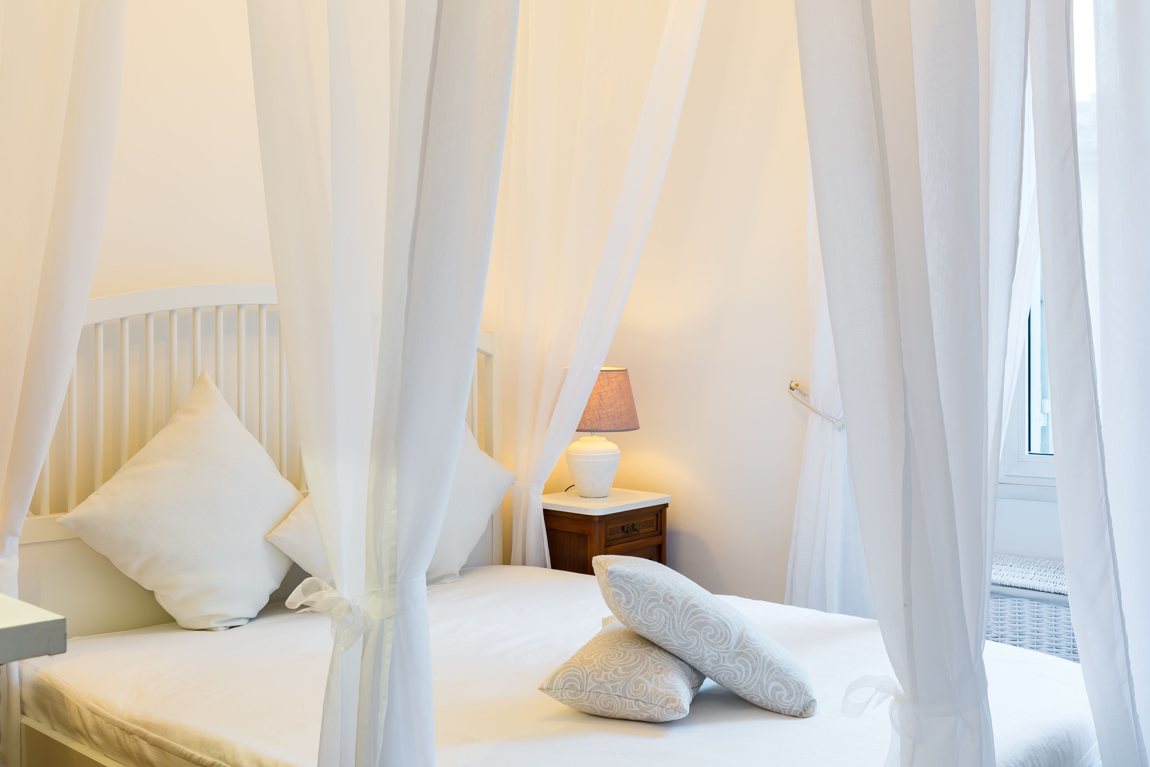 A bedroom with a canopy bed, white bedding, a bedside lamp, and a nightstand.