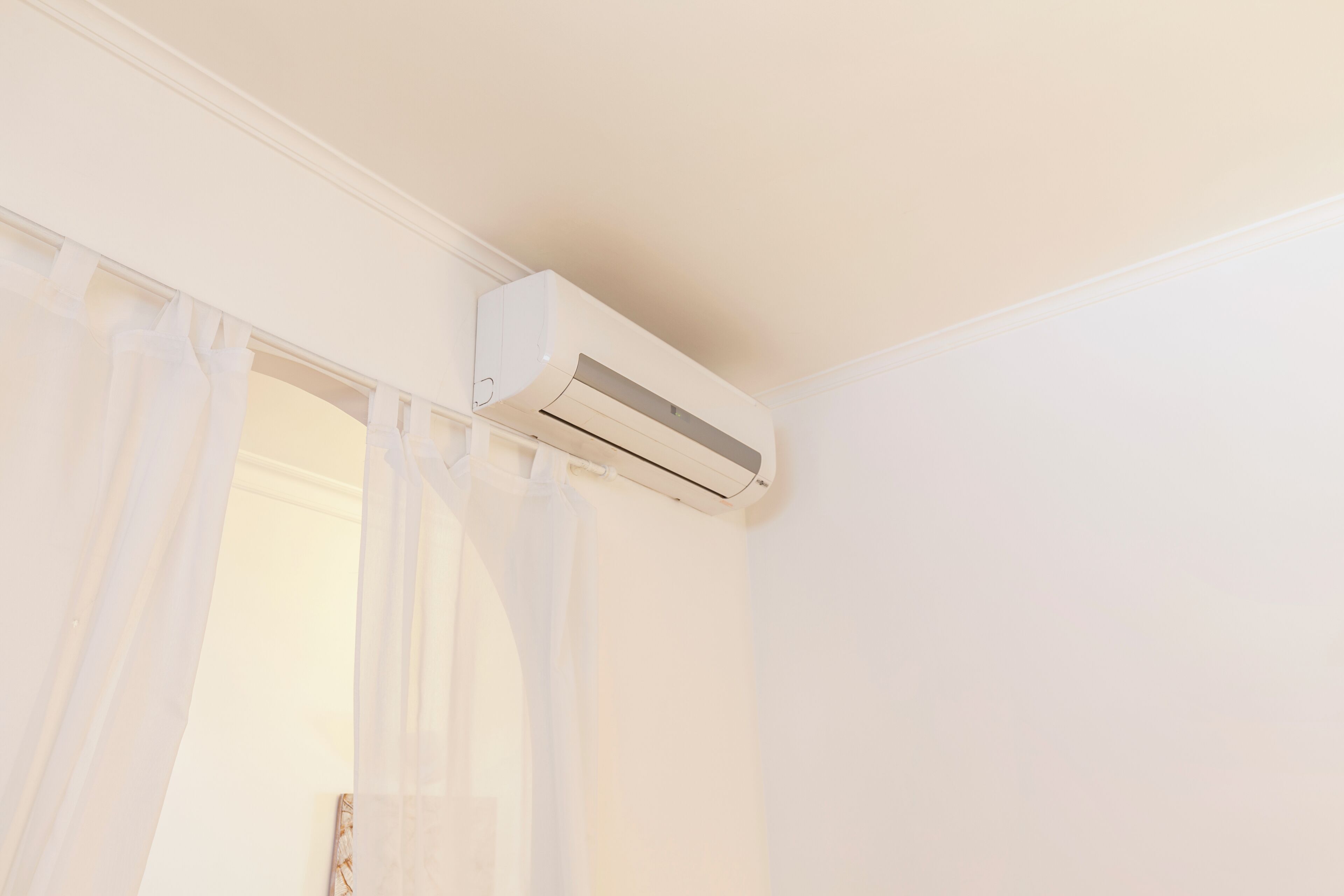 A white air conditioning unit mounted on a wall above a doorway with sheer white curtains.
