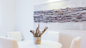A round white table with a woven basket containing sticks, surrounded by white chairs, in a room with a plain white wall and a textured artwork above. - Casa Ripetta (Rome)