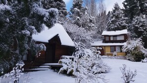 A wooden cabin with a snow-covered roof, surrounded by snow-laden trees and a clear sky.