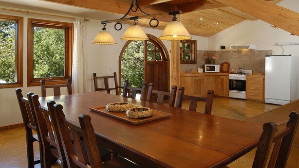 A spacious dining area with a wooden table and chairs, a kitchen with modern appliances, and large windows offering a view of greenery.