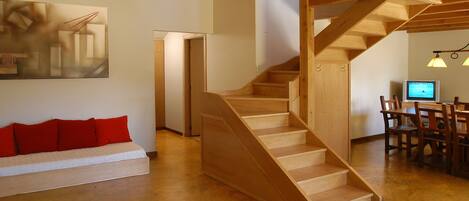 Un interior moderno con una escalera de madera, un sofá blanco con cojines rojos y un comedor con sillas de madera y un televisor.
