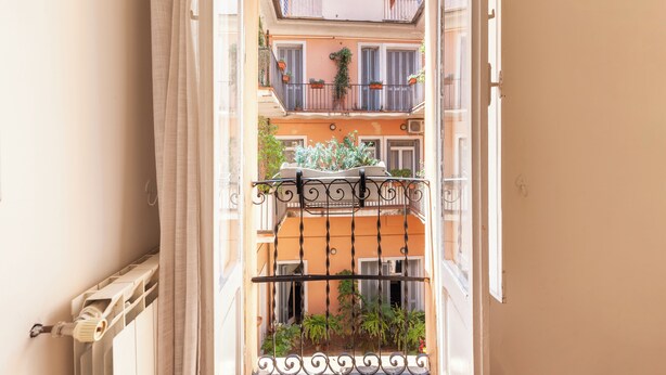 Vista a través de una ventana abierta con barandilla de metal, que muestra un edificio con varios balcones y plantas en macetas.