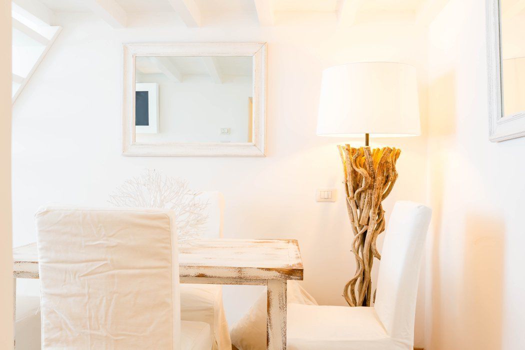 A room with a white table, two white chairs, a lamp with a twisted base, and a framed mirror on the wall.