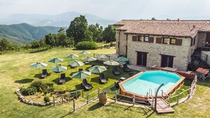 A stone building with a swimming pool, surrounded by lawn chairs and umbrellas, set against a mountainous backdrop.